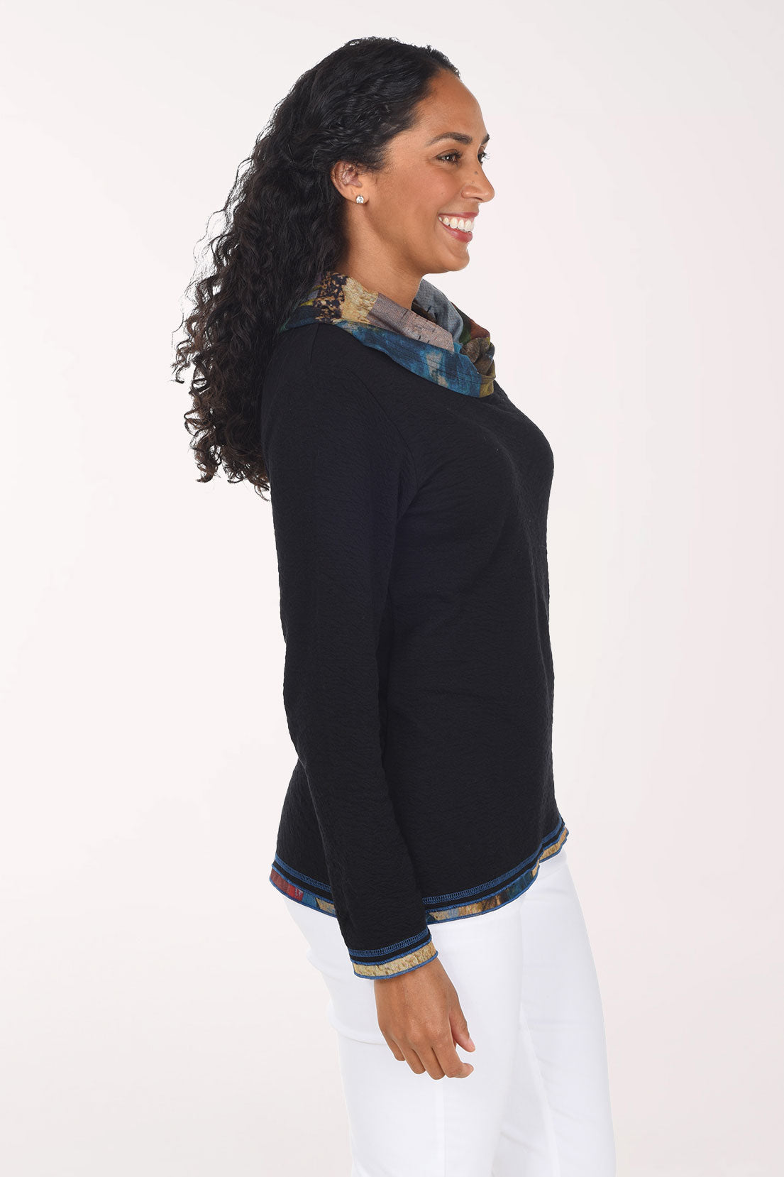 A smiling woman wearing a black long-sleeve sweater with a multicolor patterned cowl neck and hem trim, paired with white pants, against a plain white studio background.