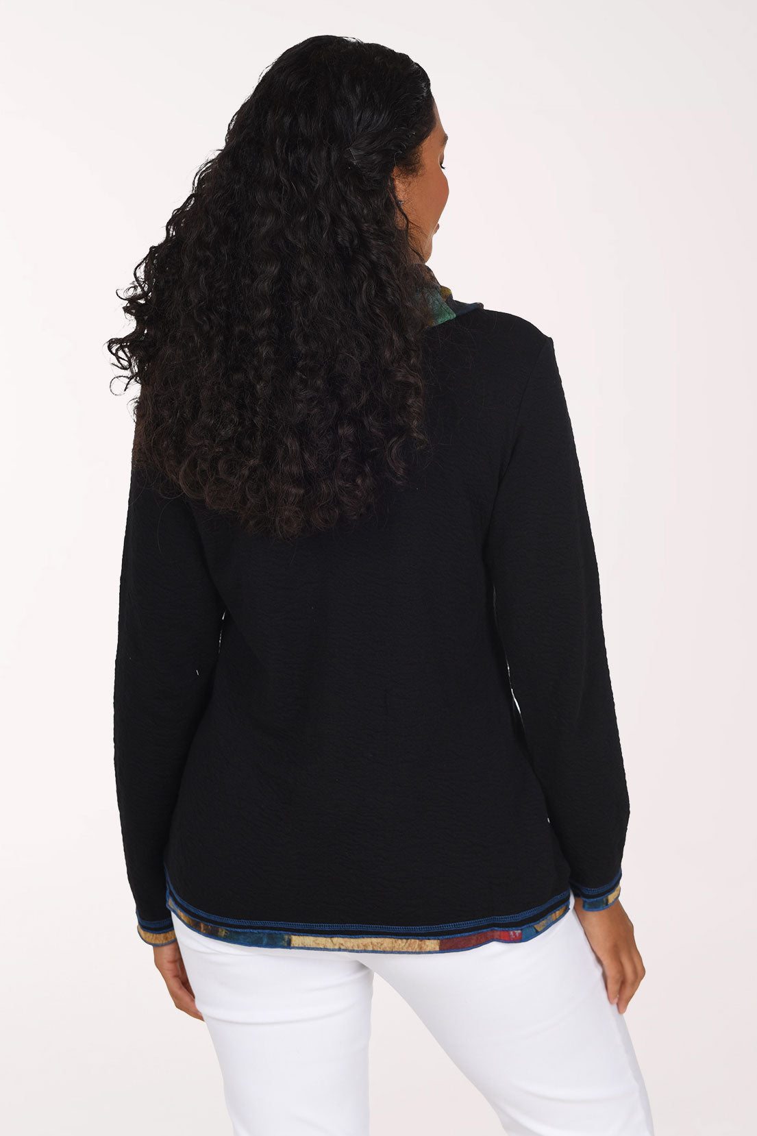 A smiling woman wearing a black long-sleeve sweater with a multicolor patterned cowl neck and hem trim, paired with white pants, against a plain white studio background.