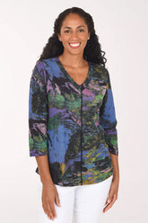 A smiling woman with long dark curly hair wearing a colorful abstract print top with three-quarter sleeves and white pants, standing against a plain white background.