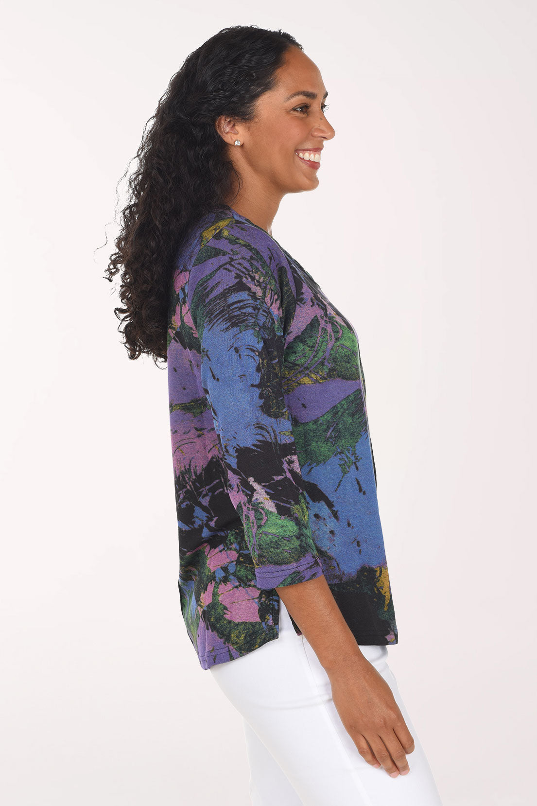 A smiling woman with long dark curly hair wearing a colorful abstract print top with three-quarter sleeves and white pants, standing against a plain white background.