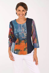 A woman modeling a three-quarter sleeve top with a multicolor abstract print on the front and sleeves, paired with white pants, against a plain white background.