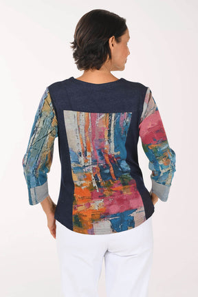 A woman shown from the back wearing a long-sleeve top with a solid navy blue upper back and shoulders, and a large multicolor abstract painted panel across the lower back and sleeves. She is wearing white pants against a plain white studio background.