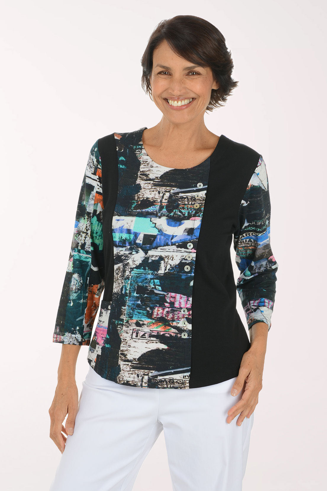 A woman modeling a three-quarter sleeve top with a black side panel and a multicolor abstract collage print on the front and sleeves, paired with white pants against a plain white background.