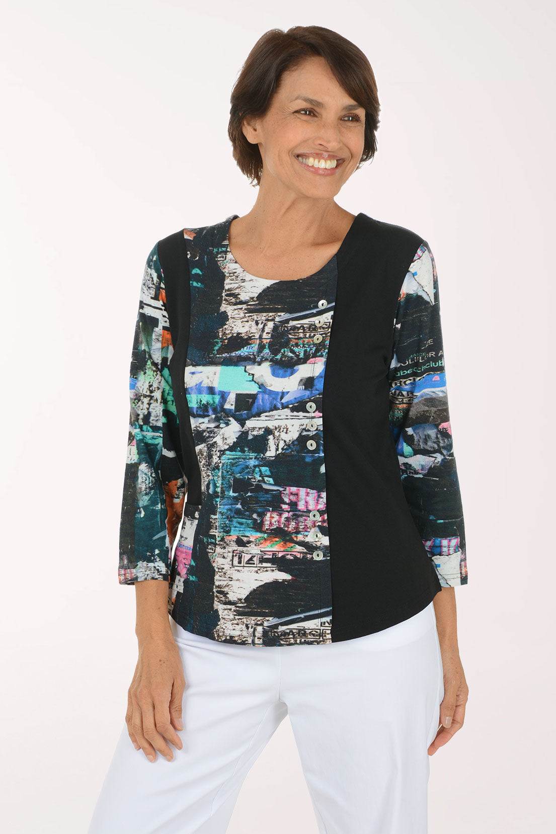 A woman modeling a three-quarter sleeve top with a black side panel and a multicolor abstract collage print on the front and sleeves, paired with white pants against a plain white background.
