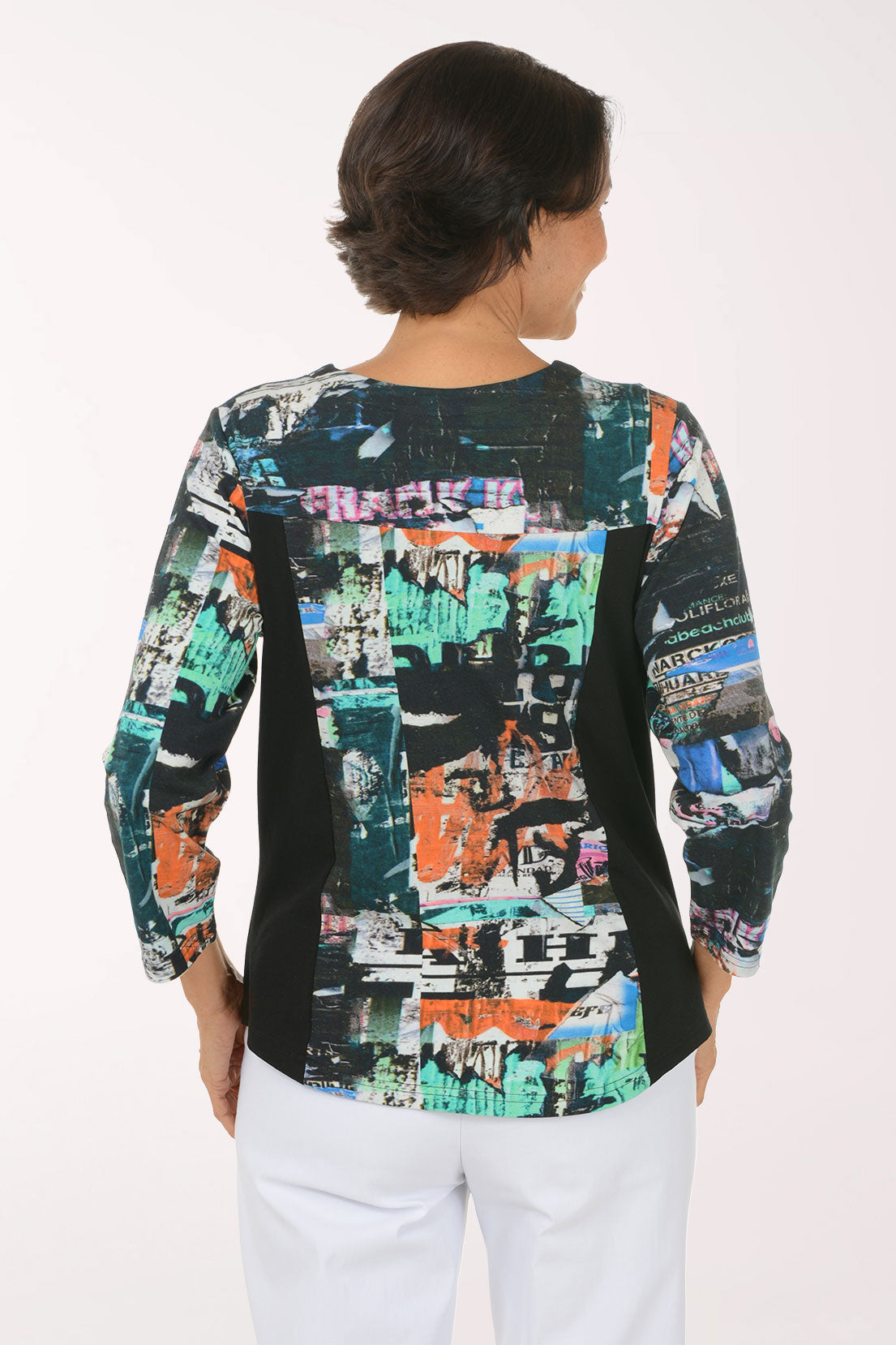 A woman modeling a three-quarter sleeve top with a black side panel and a multicolor abstract collage print on the front and sleeves, paired with white pants against a plain white background.
