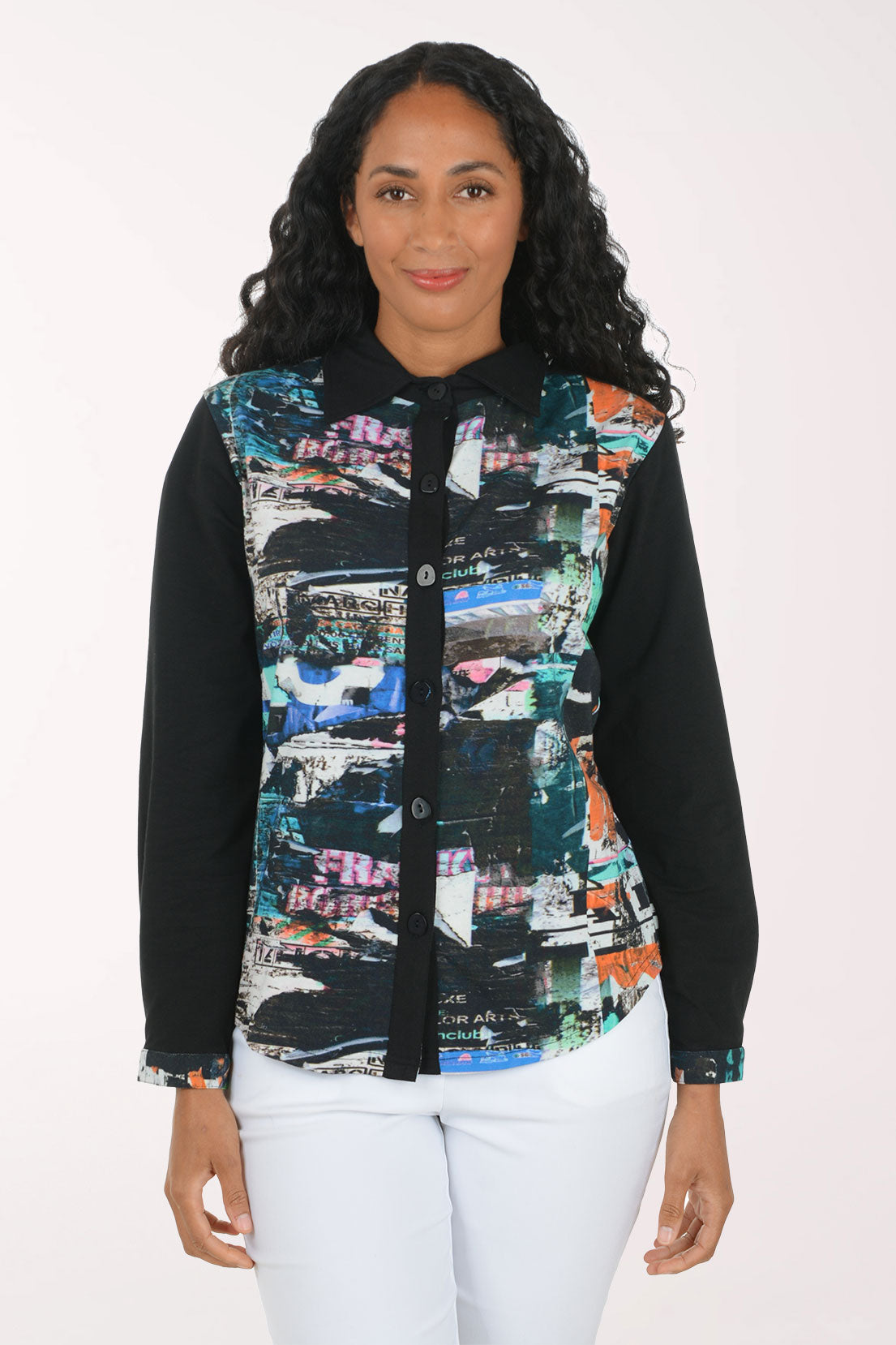 A woman modeling a long-sleeve button-up shirt with black sleeves and a colorful abstract collage print on the front panel, paired with white pants against a plain white background.