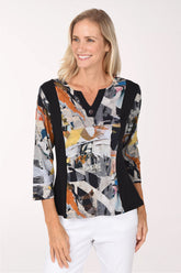 A woman wearing a long-sleeve collared blouse with a black side panel and a multicolor abstract collage print on the rest of the body, paired with white pants against a plain light background.