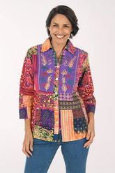 A woman modeling a vibrant, multicolored patchwork button-up shirt with intricate botanical and geometric patterns, paired with blue jeans against a plain white background.