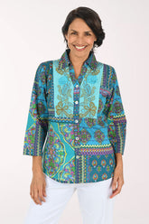A smiling woman wearing a vibrant, multicolored patchwork print blouse with three-quarter sleeves and white pants, photographed against a solid light background.