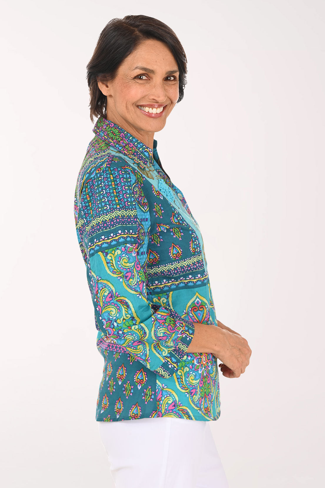 A smiling woman wearing a vibrant, multicolored patchwork print blouse with three-quarter sleeves and white pants, photographed against a solid light background.