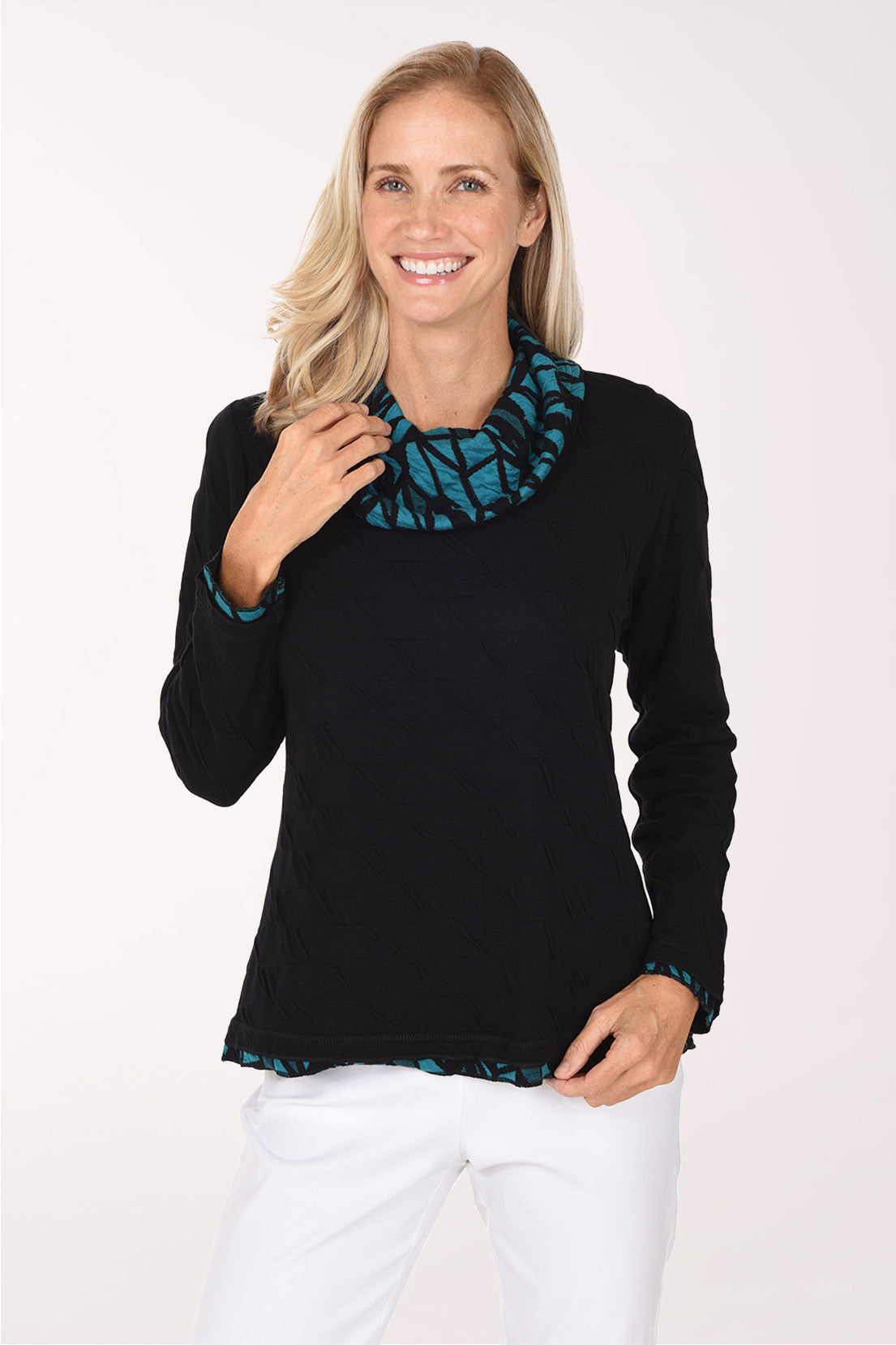 A smiling blonde woman wearing a black long-sleeve top with a teal and black patterned ruffled collar and hem, paired with white pants, against a plain white studio background.