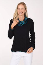 A smiling blonde woman wearing a black long-sleeve top with a teal and black patterned ruffled collar and hem, paired with white pants, against a plain white studio background.