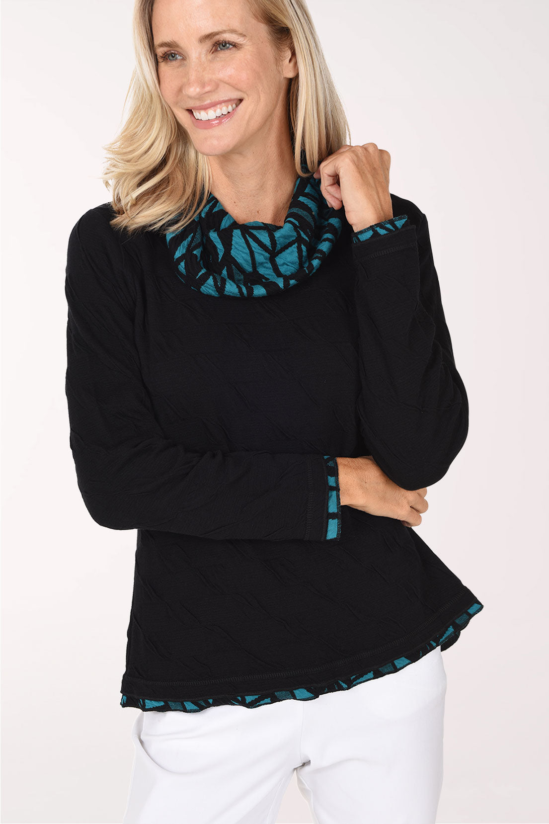 A smiling blonde woman wearing a black long-sleeve top with a teal and black patterned ruffled collar and hem, paired with white pants, against a plain white studio background.
