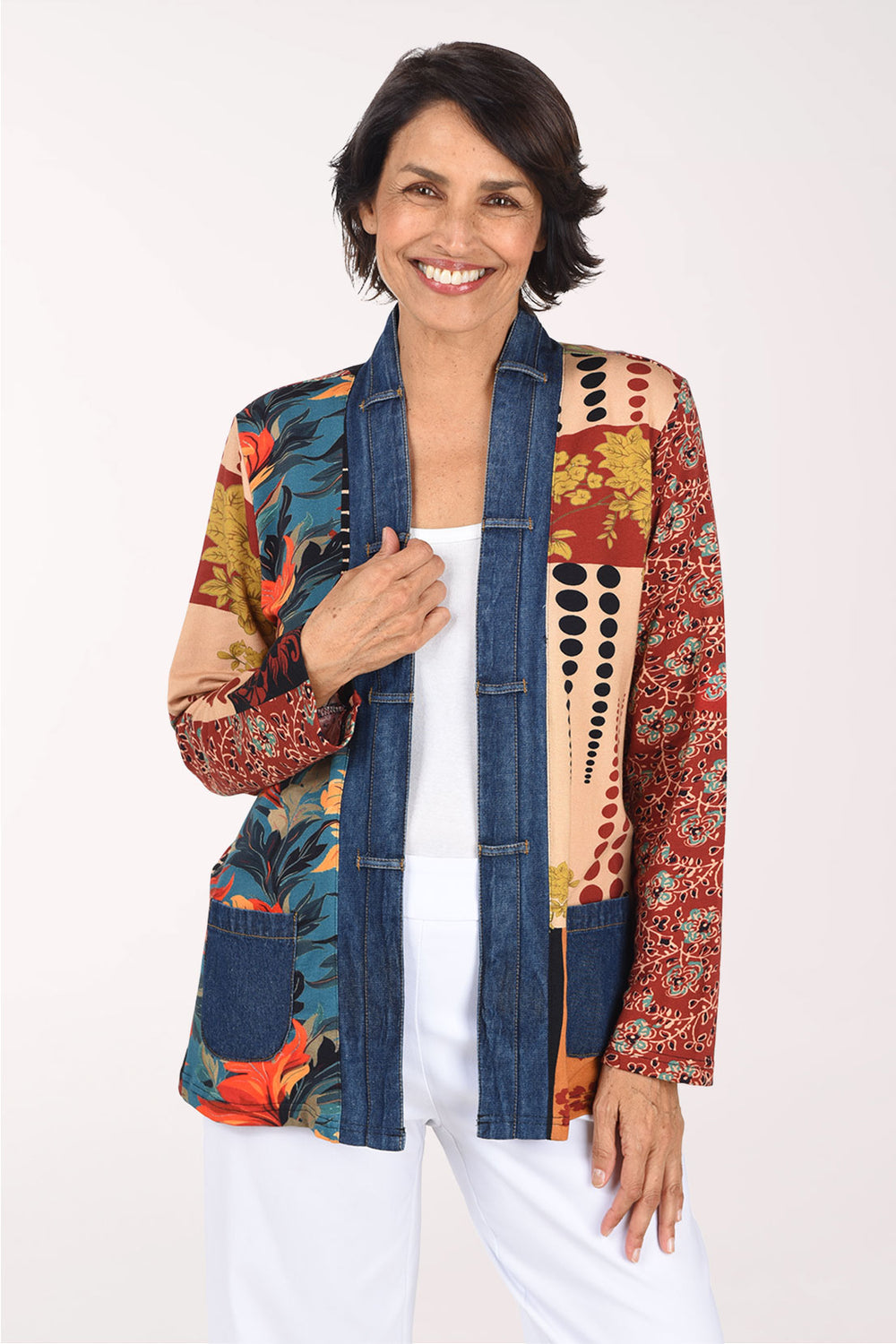 A smiling woman wearing a colorful patchwork open-front jacket with blue denim-style front panels and multicolor tropical and floral patterns on the sleeves and sides, paired with a white top and white pants, against a plain light background.