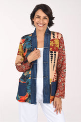 A smiling woman wearing a colorful patchwork open-front jacket with blue denim-style front panels and multicolor tropical and floral patterns on the sleeves and sides, paired with a white top and white pants, against a plain light background.