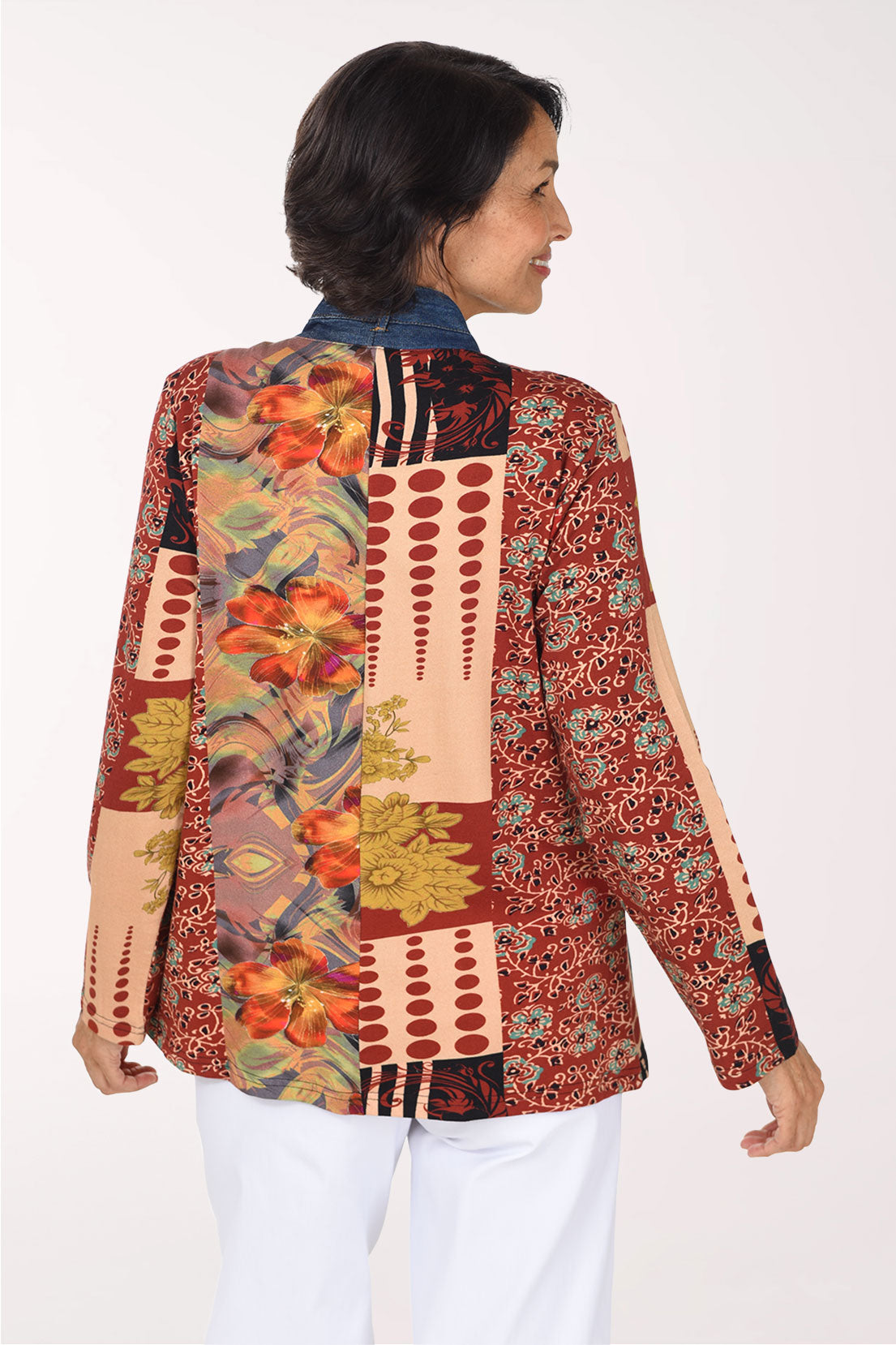 A smiling woman wearing a colorful patchwork open-front jacket with blue denim-style front panels and multicolor tropical and floral patterns on the sleeves and sides, paired with a white top and white pants, against a plain light background.