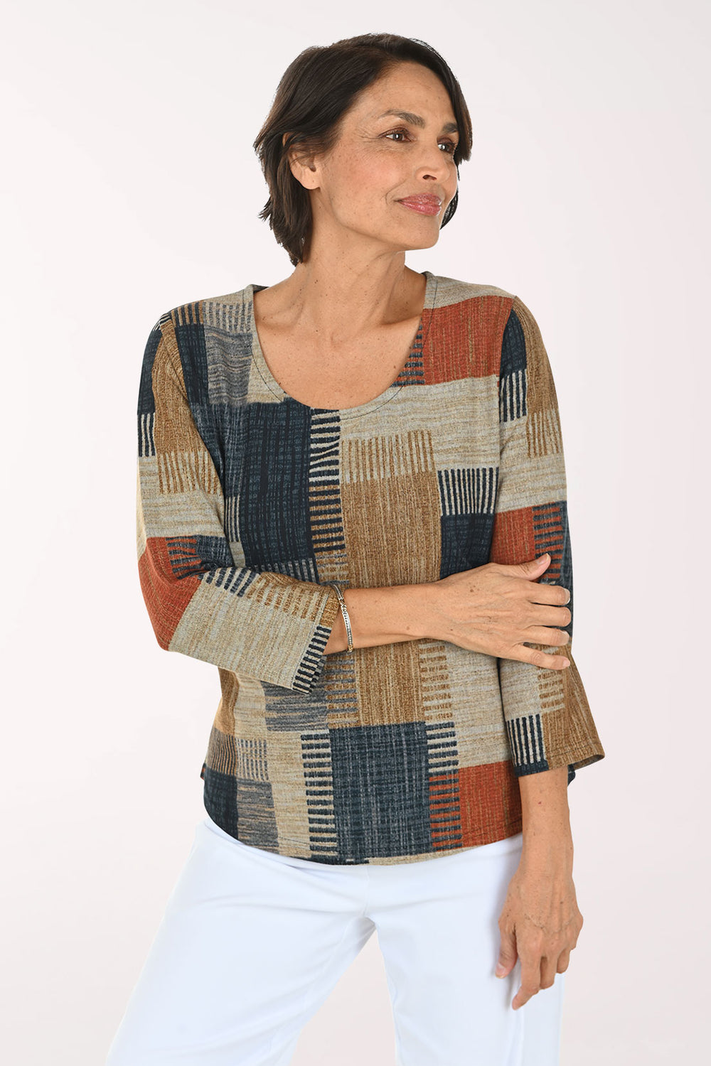 A woman modeling a three-quarter sleeve top with a patchwork geometric pattern in beige, navy, rust orange, and brown, paired with white pants against a plain white background.