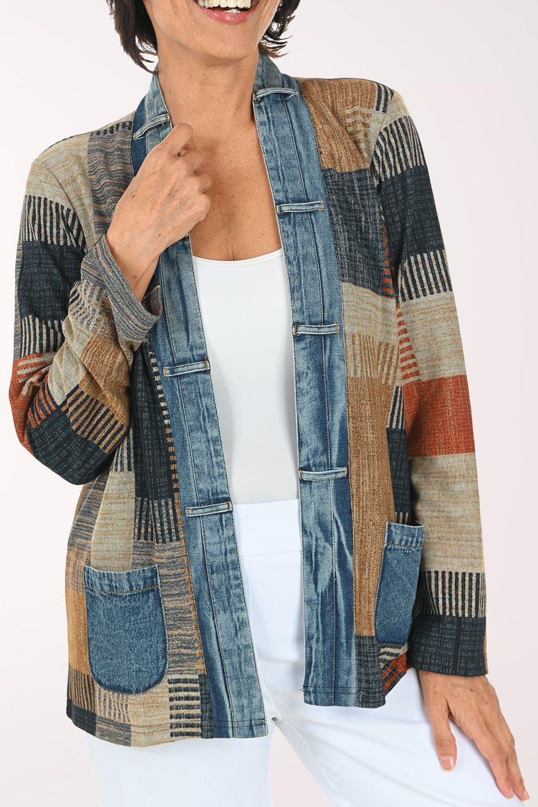 A woman modeling a patchwork cardigan with a mix of blue denim, beige, brown, and rust-orange geometric patterns, worn over a white top and white pants against a light studio background.