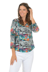 Woman wearing a colorful abstract patterned blouse with white pants on a white background.