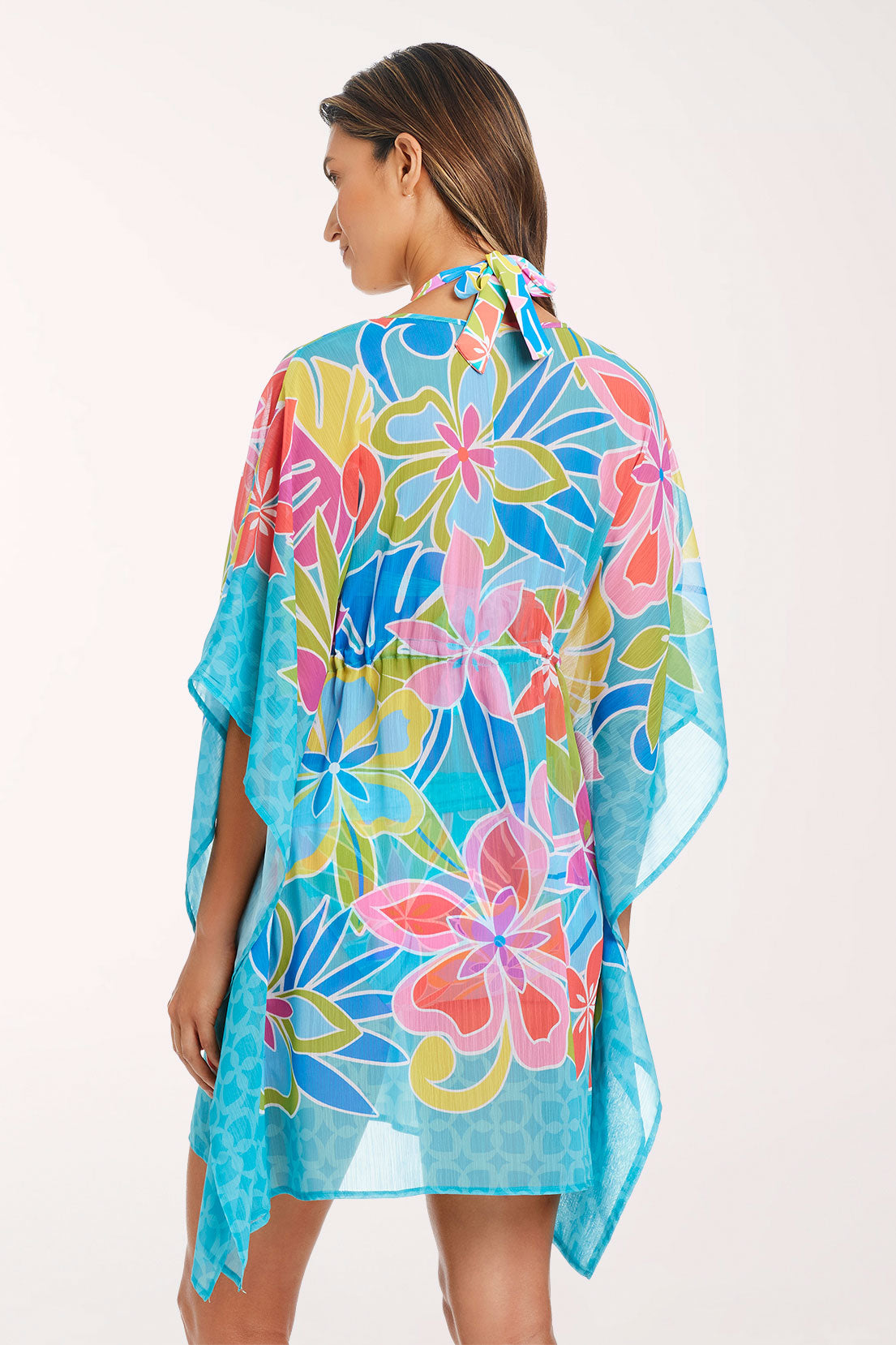 Back view of the Barcelona Bloom Chiffon Caftan, a semi-sheer turquoise tropical floral print with coral, pink, lime green and blue flowers with kimono sleeves and a turquoise geometric border at the hem, worn over a matching halter swimsuit.