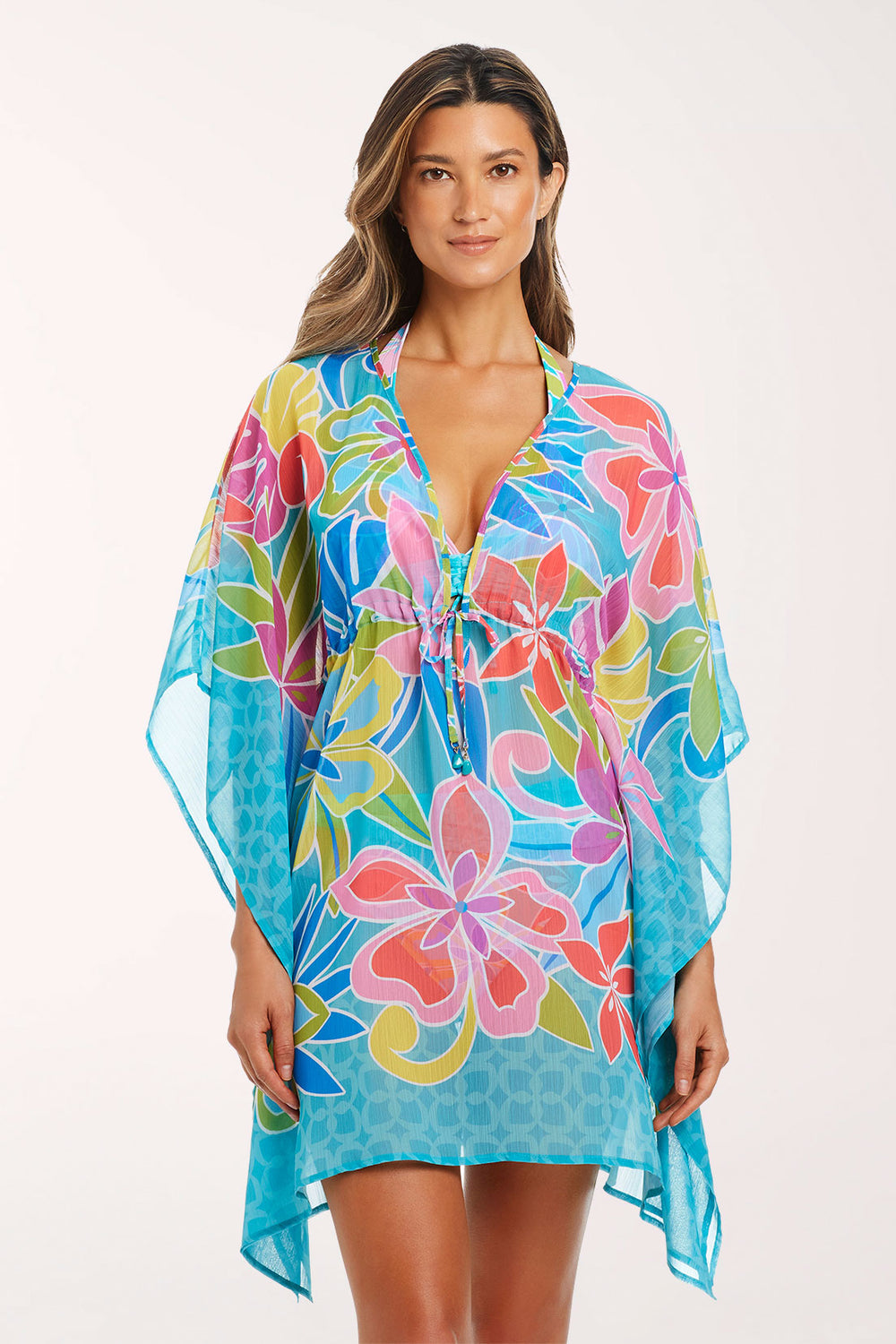 Front view of a woman wearing the Barcelona Bloom Chiffon Caftan, a semi-sheer turquoise tropical floral print with coral, pink, lime green and blue flowers, with a deep V-neck, beaded drawstring tie at the waist, wide kimono sleeves and a turquoise geometric border at the hem, over a matching swimsuit.