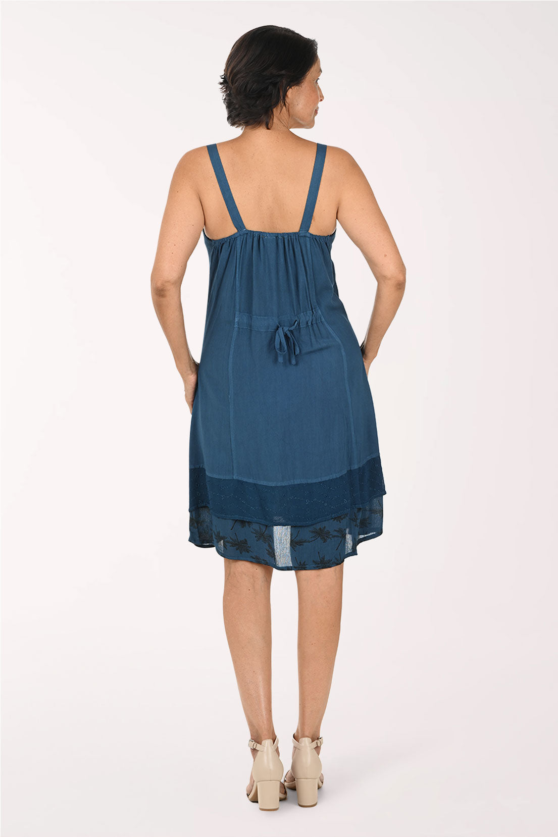 A woman modeling a sleeveless blue dress with a wrap-style bodice, front buttons, and a sheer embroidered overlay, photographed against a plain light background.