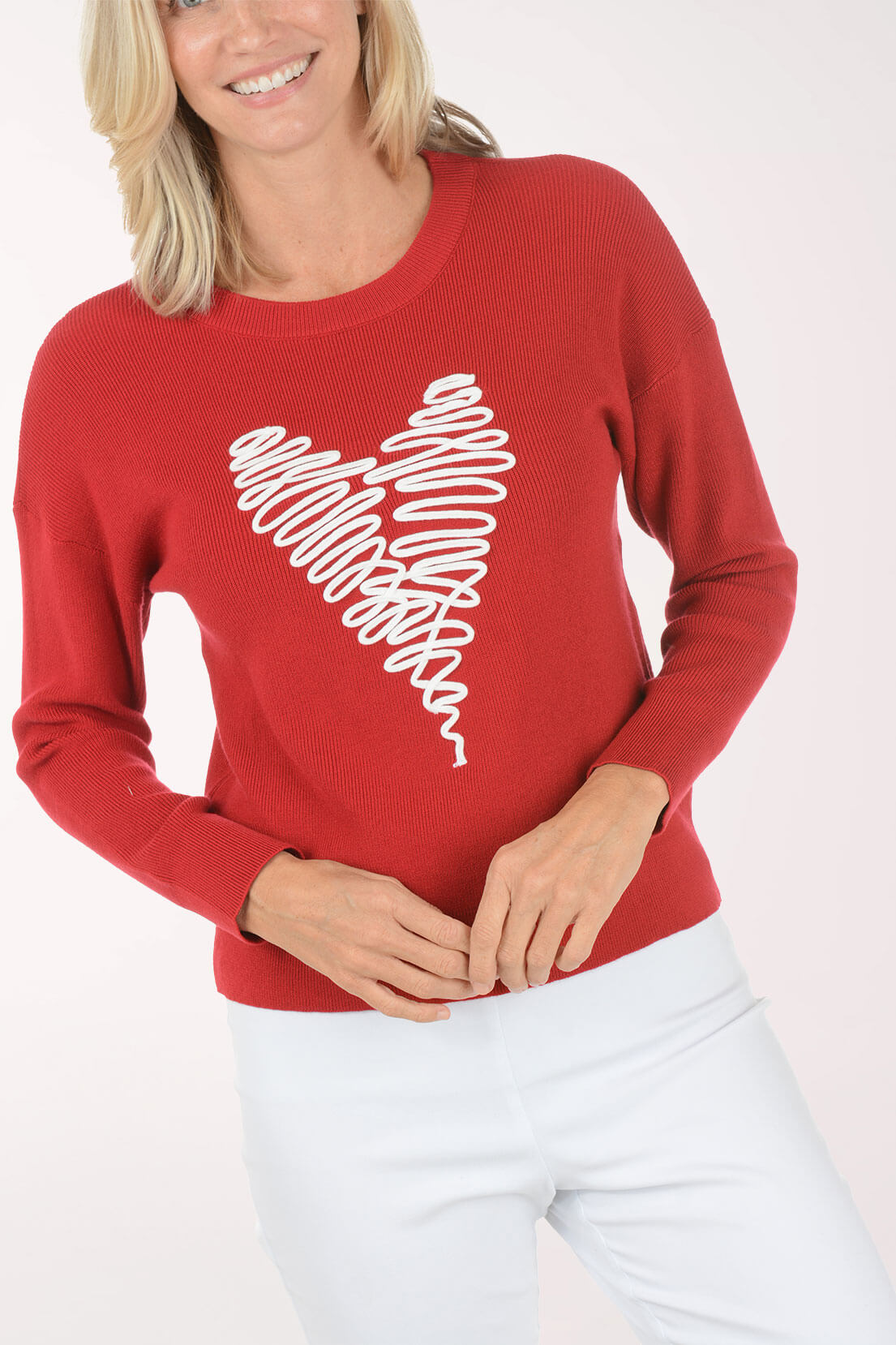 A woman wearing a red crewneck sweater with a white scribbled heart graphic on the front, paired with white pants, against a plain white background.