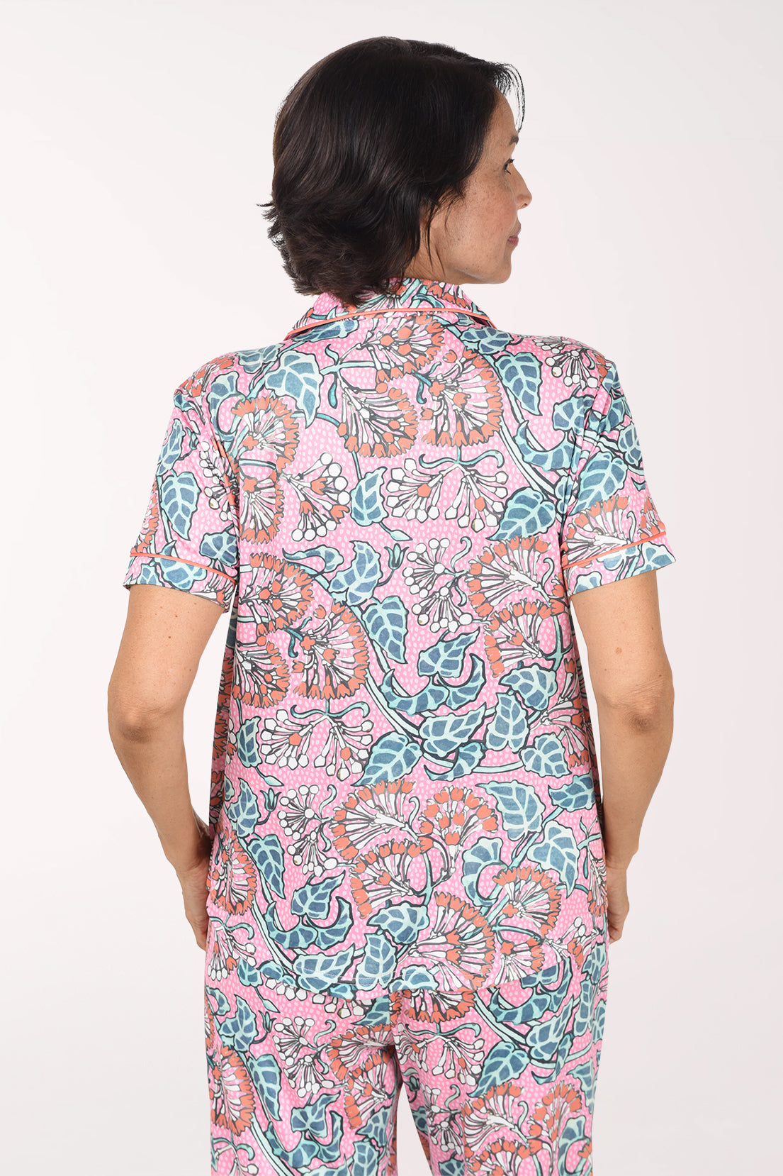 A woman shown from the back wearing a short-sleeve collared pajama top and matching pants set with a pink base and a teal, orange, and black paisley pattern, against a plain white background.