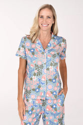 A smiling blonde woman wearing a short-sleeve pajama set with a light blue background and a colorful floral print, standing against a plain white studio background.