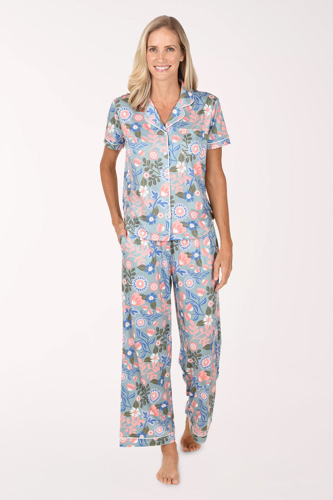 A woman modeling a two-piece short-sleeve pajama set with a colorful floral and paisley print on a light blue background, set against a plain white studio background.