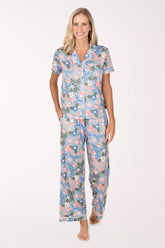 A woman modeling a two-piece short-sleeve pajama set with a colorful floral and paisley print on a light blue background, set against a plain white studio background.