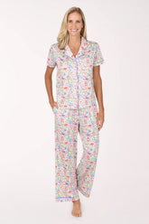 A woman modeling a matching short-sleeve button-up pajama shirt and wide-leg pajama pants set with a colorful tropical print on a white background.