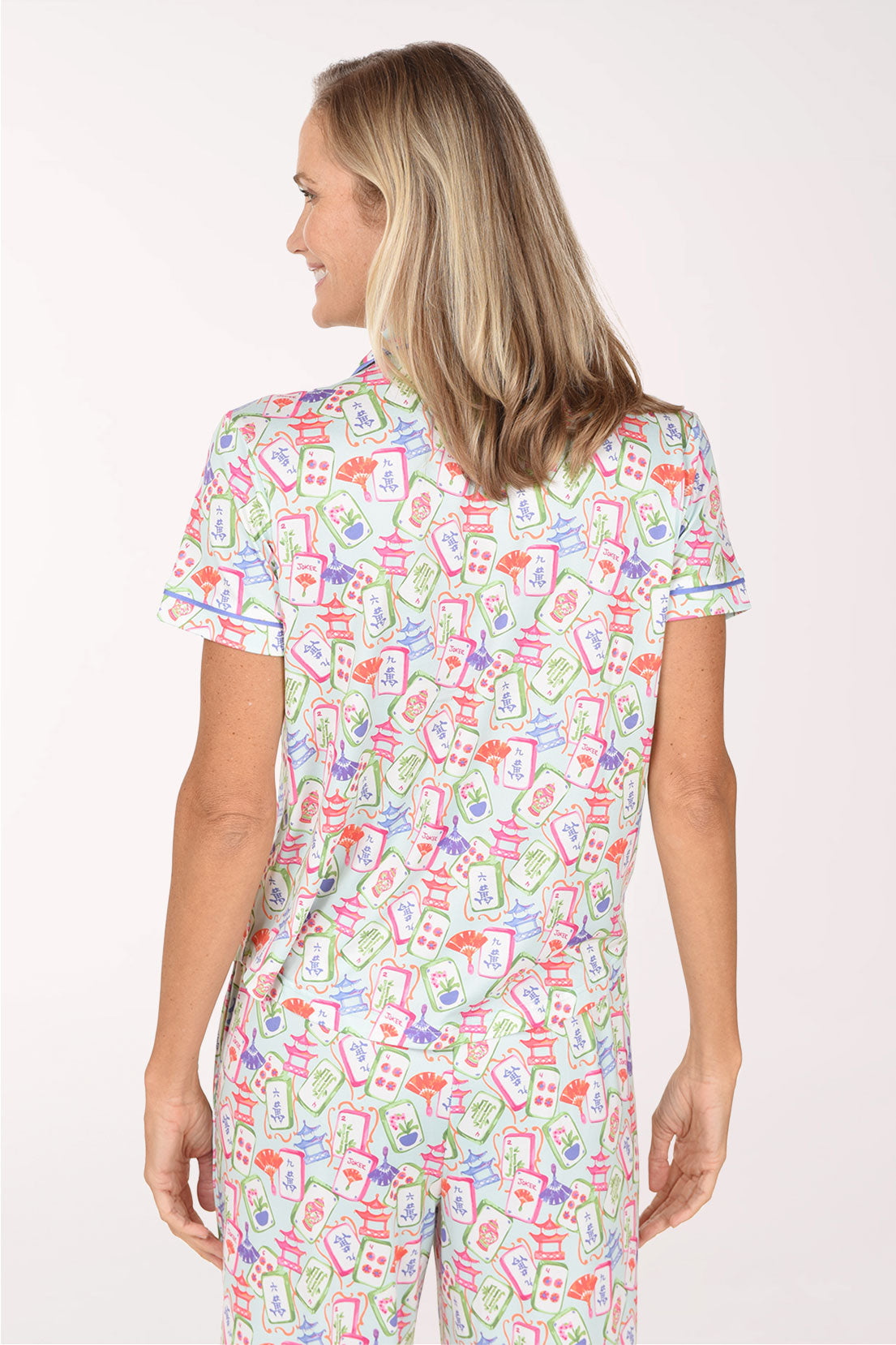 A smiling blonde woman wearing a short-sleeve button-front pajama top and matching pants with a colorful cartoon-style print on a white base, photographed against a plain light background.