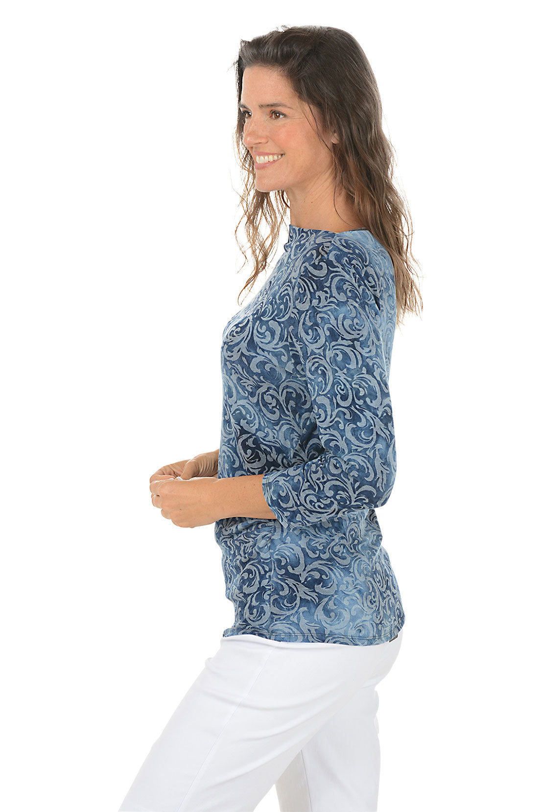 Woman wearing a 3/4 sleeve blue tie-dye indigo scroll patterned top and white pants on a white background. Side shot.
