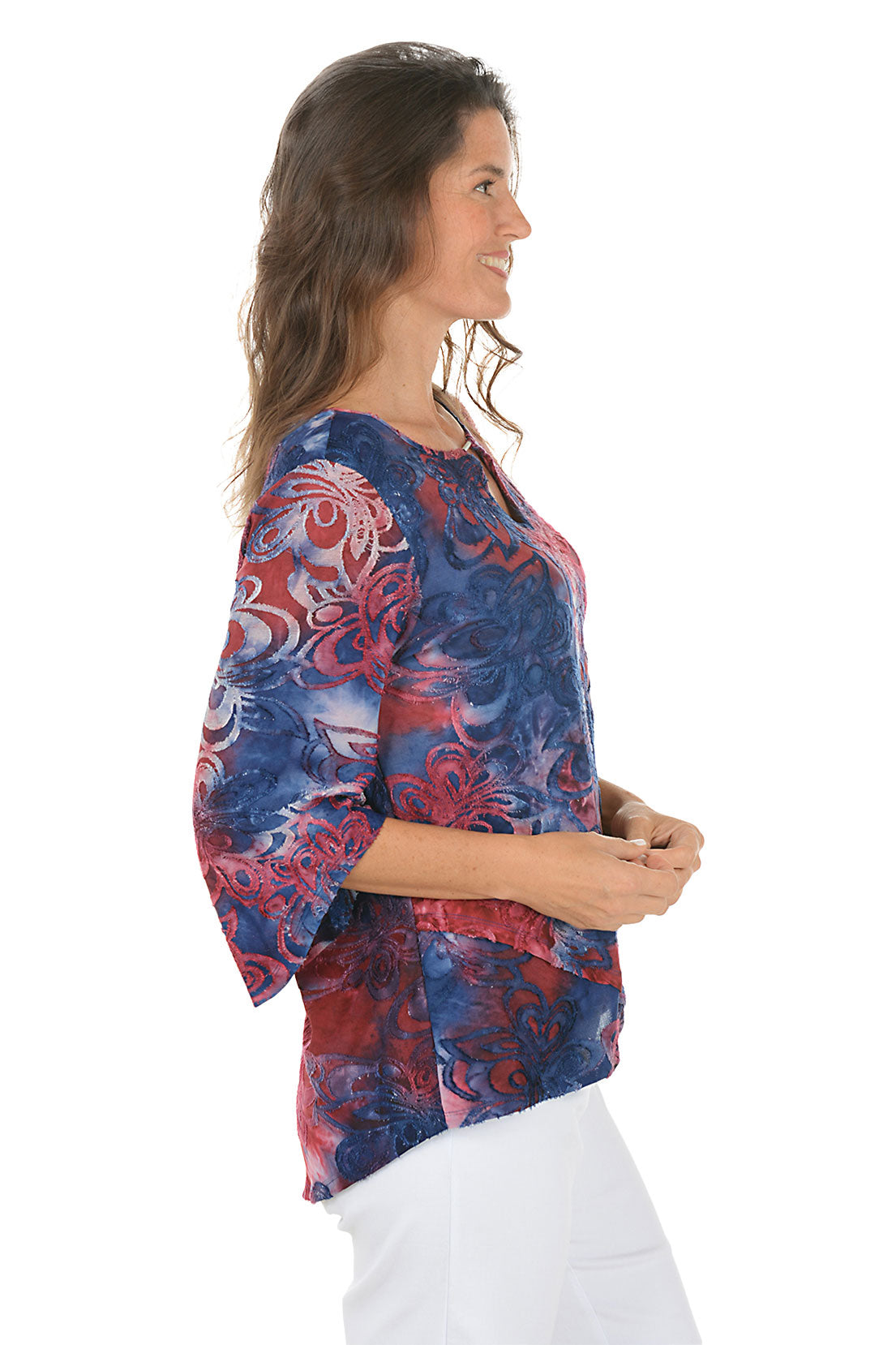 Woman wearing a colorful red and blue floral blouse with white pants on a white background. Side shot.