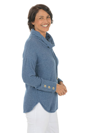 Woman wearing a blue cowl-neck sweater with white pants on a white background. Side shot.
