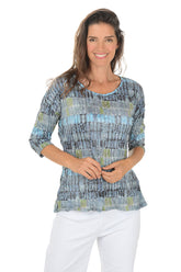Woman wearing a patterned blue and gray top with white pants on a white background.