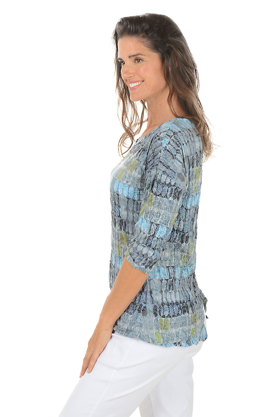 Woman wearing a patterned blue and gray top with white pants on a white background. Side shot.