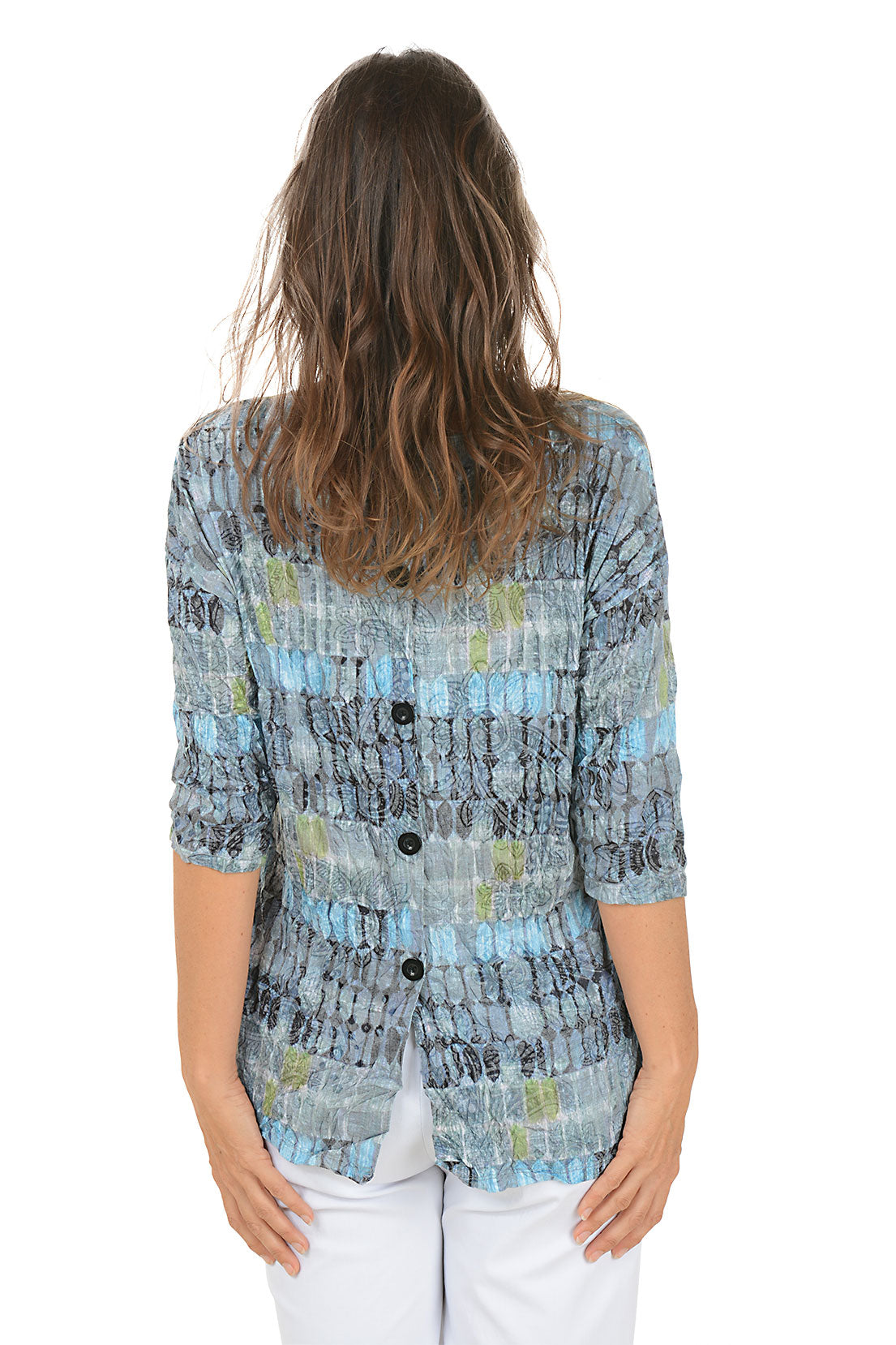 Woman wearing a patterned blue and gray top with white pants on a white background. Back shot.