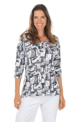 Woman wearing a grey geometric patterned top and white pants on a white background.