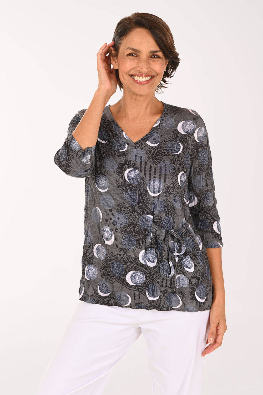 A smiling woman wearing a dark gray wrap-style blouse with a moon phase pattern and white pants, photographed against a plain white studio background.