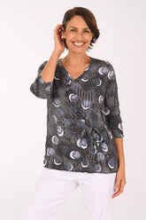 A smiling woman wearing a dark gray wrap-style blouse with a moon phase pattern and white pants, photographed against a plain white studio background.
