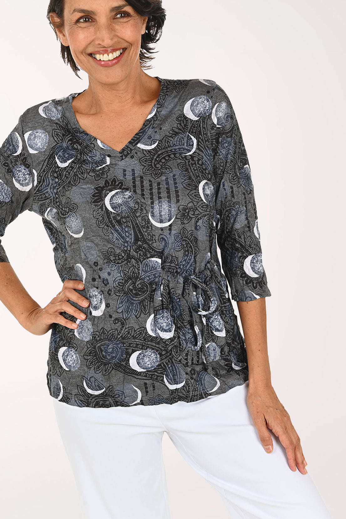 A smiling woman wearing a dark gray wrap-style blouse with a moon phase pattern and white pants, photographed against a plain white studio background.