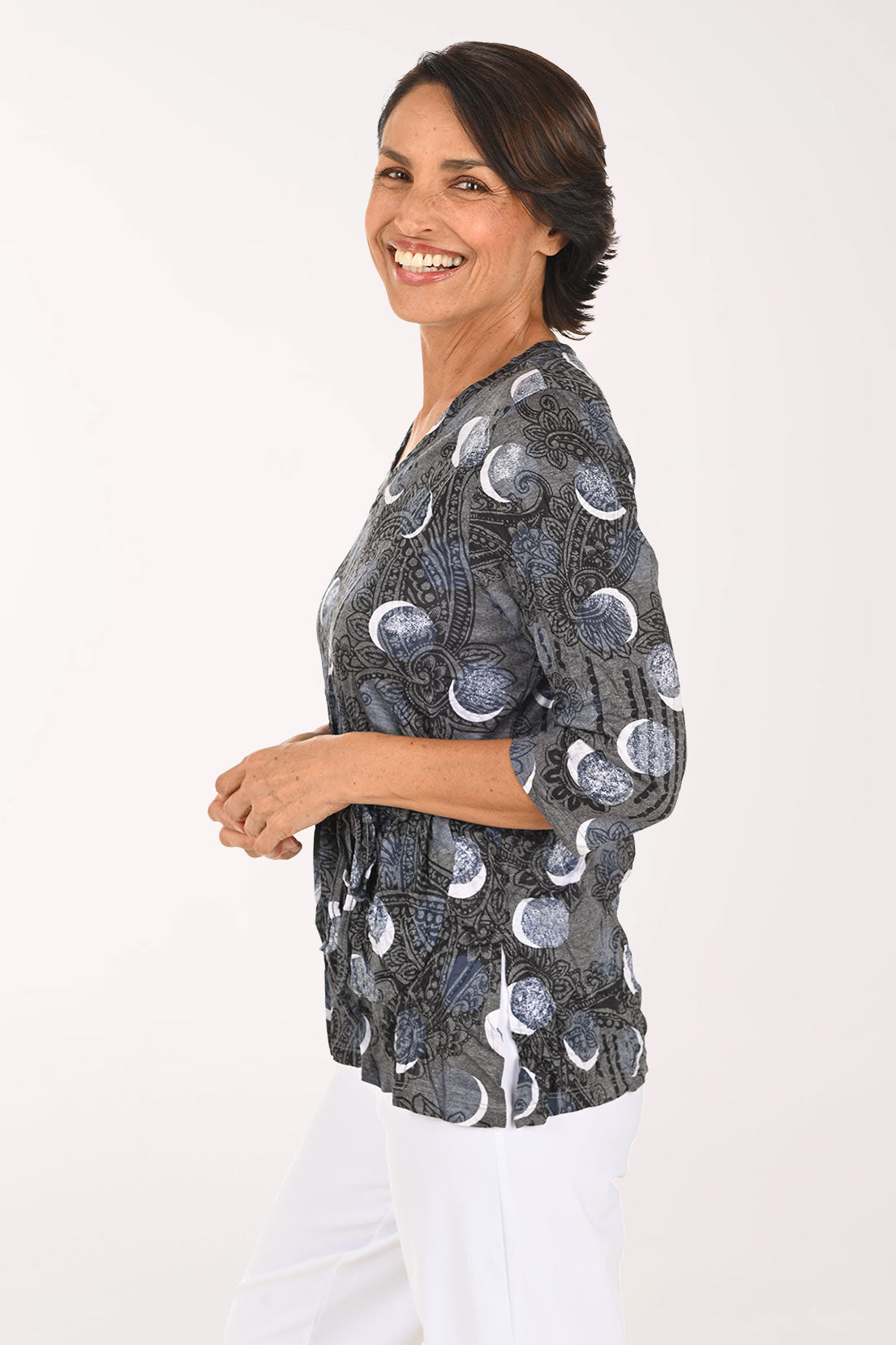 A smiling woman wearing a dark gray wrap-style blouse with a moon phase pattern and white pants, photographed against a plain white studio background.