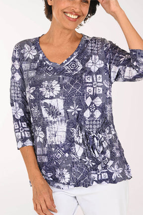 A woman wearing a navy blue and white patterned three-quarter sleeve top with a V-neck and gathered waist, paired with white pants, against a plain white background.