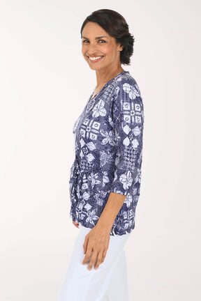 A woman wearing a navy blue and white patterned blouse with three-quarter sleeves, paired with white pants, photographed against a plain white studio background.