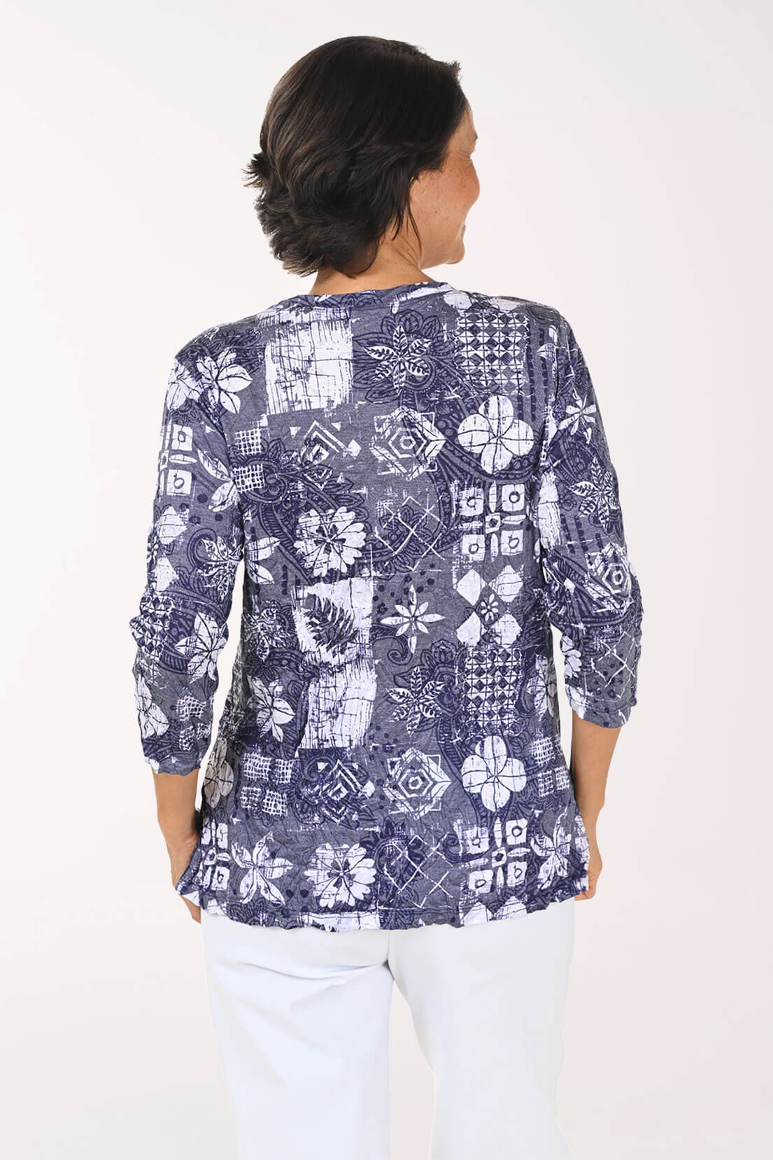 A woman viewed from the back wearing a navy blue and white patterned blouse with three-quarter sleeves and white pants, set against a plain white studio background.