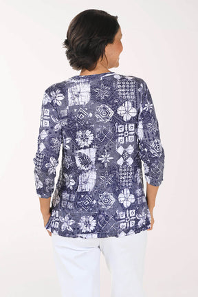 A woman viewed from the back wearing a navy blue and white patterned blouse with three-quarter sleeves and white pants, set against a plain white studio background.
