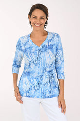 A smiling woman wearing a blue and white tie-dye patterned wrap top with three-quarter sleeves and white pants, photographed against a plain light background.