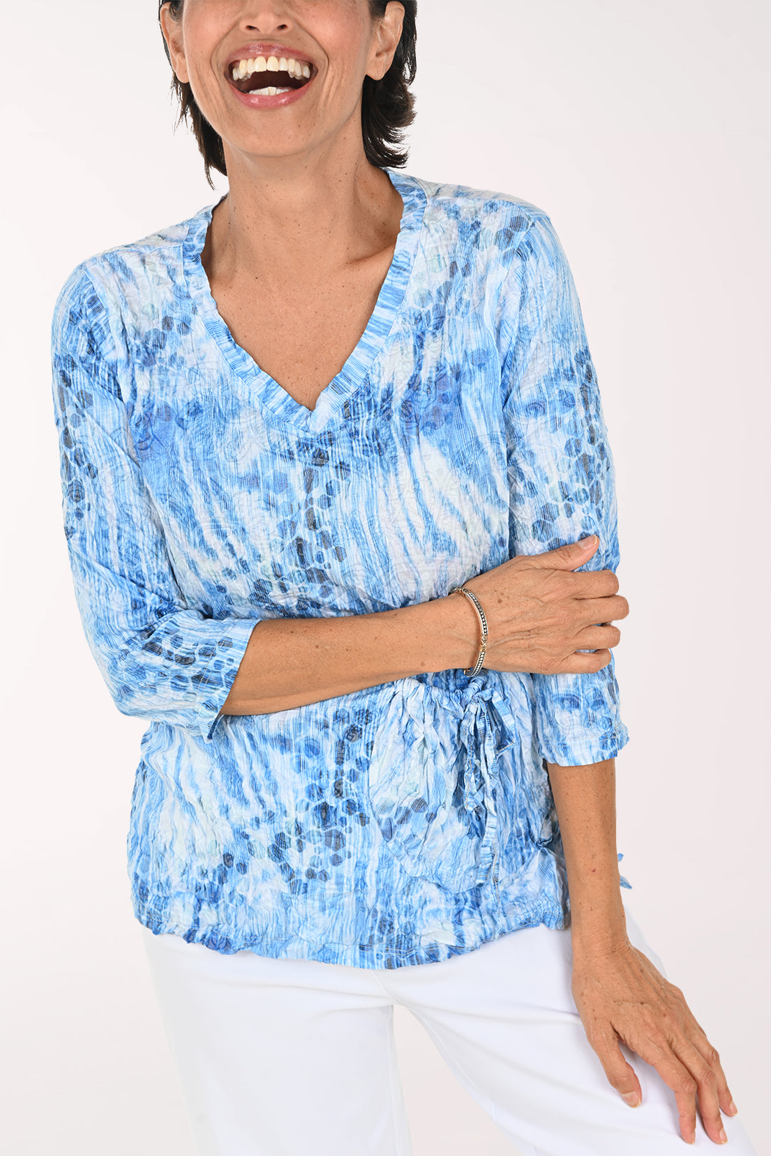 A smiling woman wearing a blue and white tie-dye patterned wrap top with three-quarter sleeves and white pants, photographed against a plain light background.